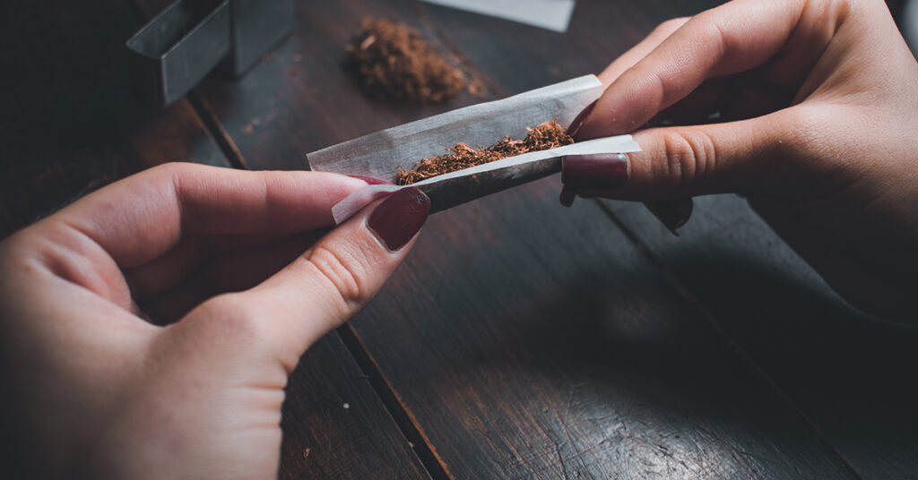 A close-up of a person's hands carefully rolling loose tobacco into a cigarette using rolling paper over a wooden table.