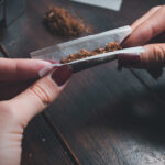 A close-up of a person's hands carefully rolling loose tobacco into a cigarette using rolling paper over a wooden table.