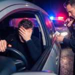 A police officer shining a flashlight into a car while inspecting the driver's license of a distressed man holding a beer bottle at night.