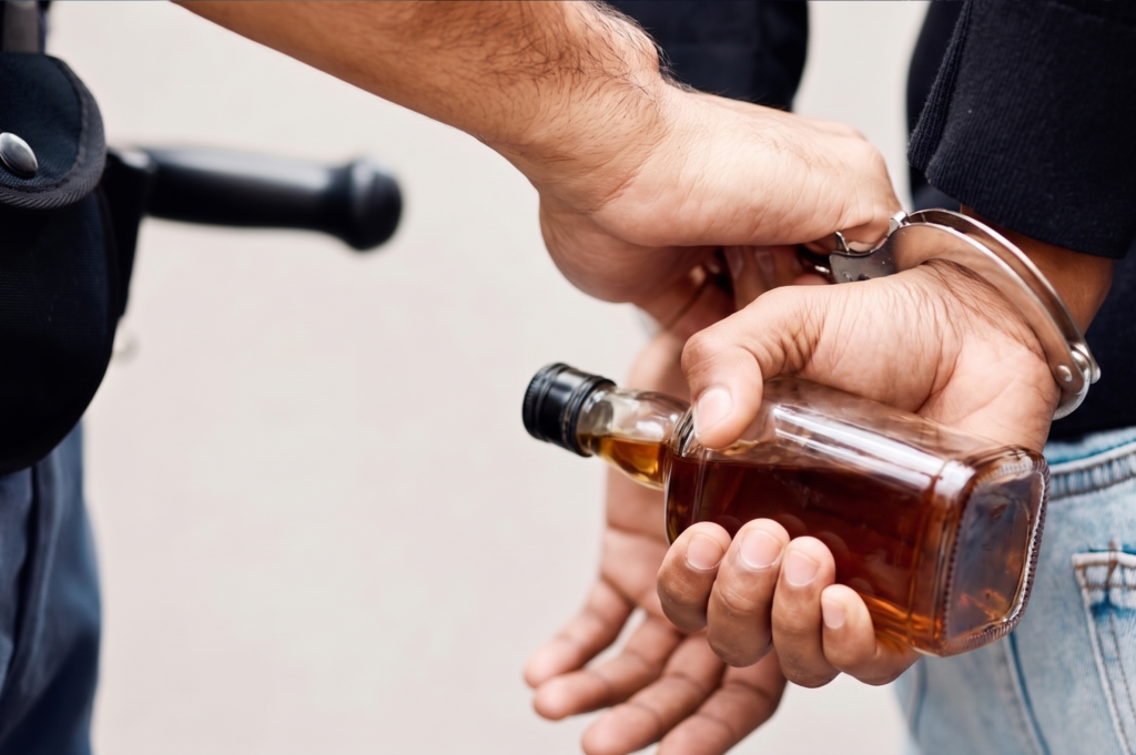 Close-up of a police officer putting handcuffs on a person who is holding a small glass liquor bottle behind their back.