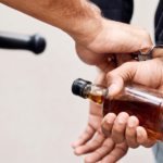 Close-up of a police officer putting handcuffs on a person who is holding a small glass liquor bottle behind their back.