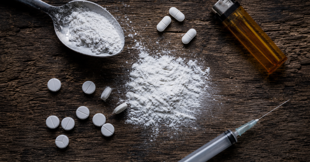 Drug paraphernalia with pills, powder, syringe, and lighter on a wooden table.