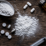 Drug paraphernalia with pills, powder, syringe, and lighter on a wooden table.
