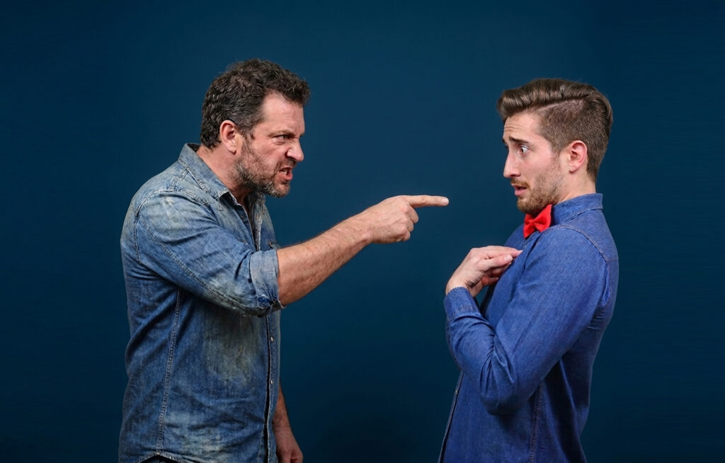 An angry man in a denim shirt aggressively points his finger at a nervous-looking younger man wearing a red bowtie, who is pointing at himself in surprise.