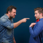 An angry man in a denim shirt aggressively points his finger at a nervous-looking younger man wearing a red bowtie, who is pointing at himself in surprise.