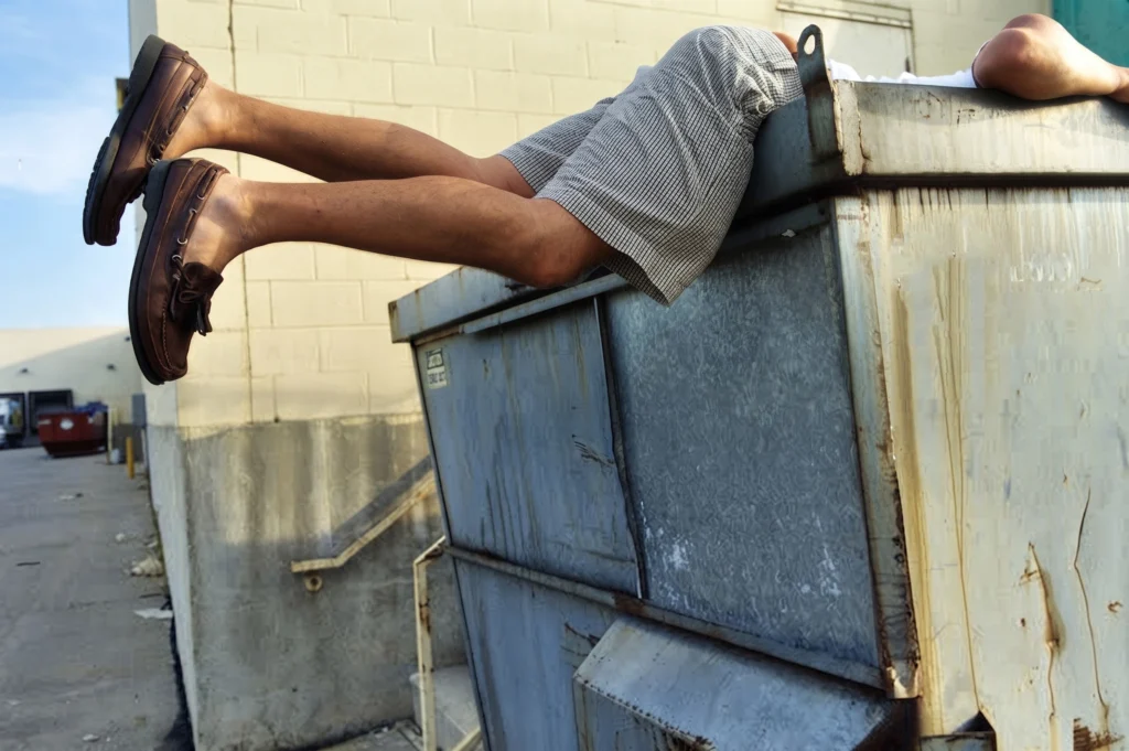 A person in shorts and brown loafers leaning headfirst inside a large outdoor trash dumpster.