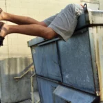 A person in shorts and brown loafers leaning headfirst inside a large outdoor trash dumpster.