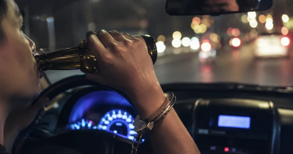 Driver drinking from a bottle at night while holding the wheel, with a handcuff on the wrist and blurred city lights ahead.