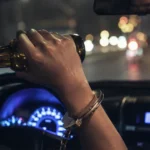 Driver drinking from a bottle at night while holding the wheel, with a handcuff on the wrist and blurred city lights ahead.