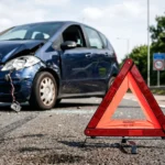 A red warning triangle stands on foreground, with a badly damaged blue car and a police officer taking notes.