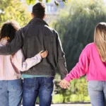 Rear view of a man walking with his arm around one woman while holding hands with another woman beside him.
