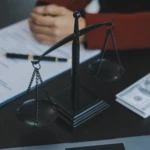 Black scales of justice on a desk with legal papers and a stack of money in the background.