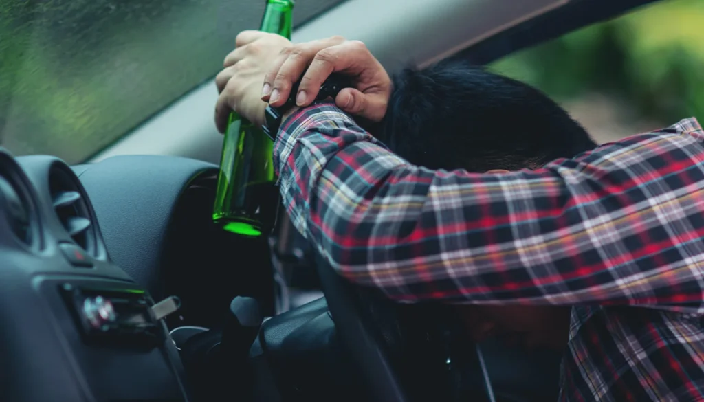 Drunk driver leaning on the steering wheel holding a beer bottle.