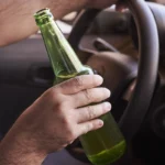Driver holding a green beer bottle while sitting behind steering wheel.