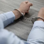 Close-up of a person's handcuffed hands resting on a wooden table.
