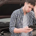 Frustrated man looking at his smartphone in front of an open car hood