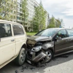 Two cars involved in a traffic collision on a street, showing severe front-end damage on a brown hatchback that crashed into the back of a cream-colored vehicle