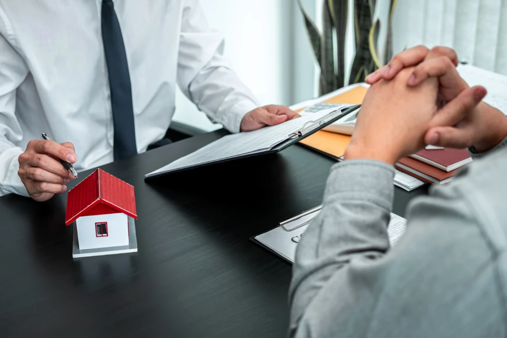 A small model house with a red roof sitting on a desk during a consultation between a professional holding a clipboard and a client