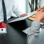 A small model house with a red roof sitting on a desk during a consultation between a professional holding a clipboard and a client