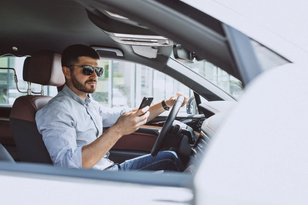 A man wearing sunglasses holding a smartphone while sitting in the driver's seat of a vehicle with a hand on the steering wheel.